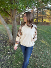 Load image into Gallery viewer, Woman standing outdoors in a park wearing a cream sweater with patterned sleeves and blue jeans.