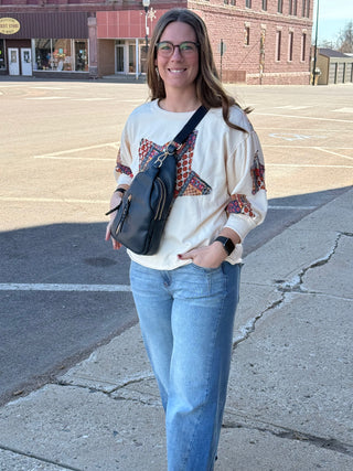 Woman standing on a street corner wearing a white sweater with colorful patterns and blue jeans, holding a black bag.
