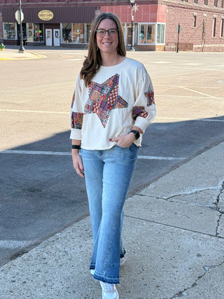 Woman standing on a street corner wearing a white sweater with star patterns and blue jeans.