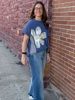 Woman wearing a blue t-shirt with a colorful design and jeans, standing against a brick wall.
