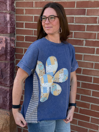Woman wearing a blue t-shirt with a floral patch design against a brick wall.