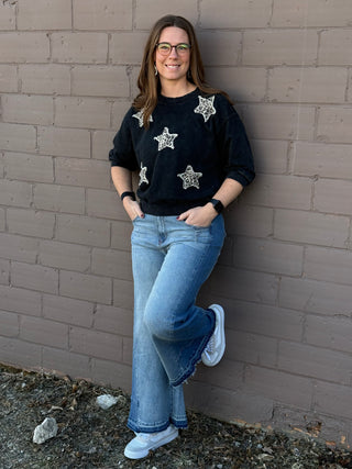 Person wearing a black sweater with star patterns and blue jeans leaning against a brick wall.