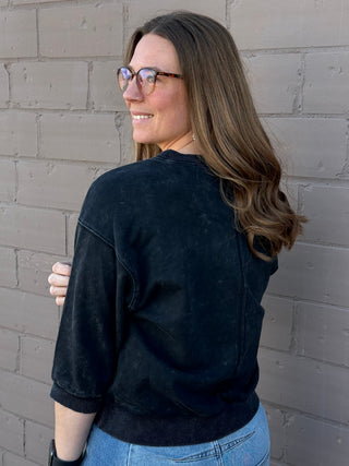 Woman wearing a black sweater and blue jeans standing against a gray brick wall.