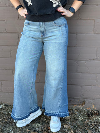 Person wearing blue flared jeans and white sneakers against a brick wall.