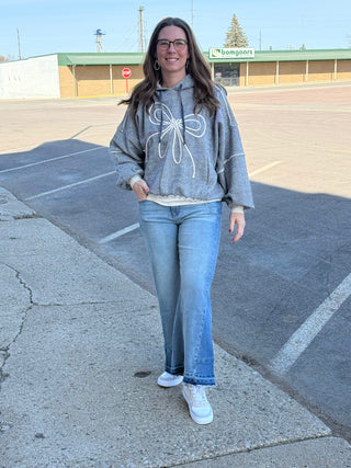 Woman wearing a gray sweatshirt with a bow design and blue jeans standing on a sidewalk.