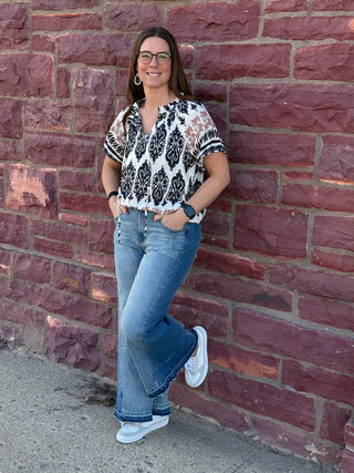 Woman standing against a brick wall wearing a patterned blouse and jeans.