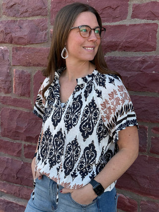 Woman wearing a patterned blouse and jeans standing against a brick wall.