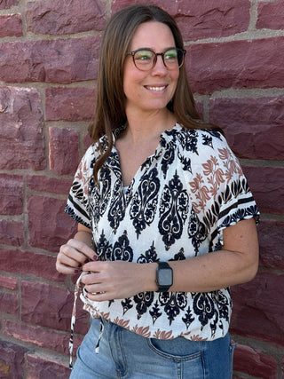 Woman wearing a patterned blouse and glasses standing against a brick wall.