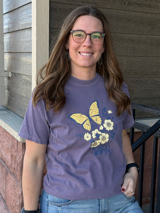 Woman wearing a purple t-shirt with butterfly and flower design standing against a wooden wall.