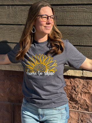 Woman wearing a gray t-shirt with a sunflower graphic and 'choose to shine' text, standing against a wooden wall.