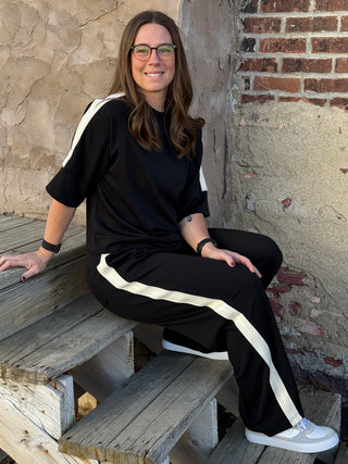 Woman sitting on a wooden bench wearing a black outfit with white stripes, against a stone wall.