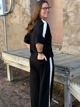 Woman wearing a black outfit with white stripes standing outdoors near a wooden bench.
