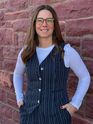 Woman wearing a blue striped vest over a white long-sleeve shirt against a brick wall.