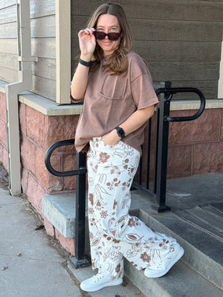 Woman standing on steps wearing a brown oversized shirt and patterned pants, holding sunglasses.