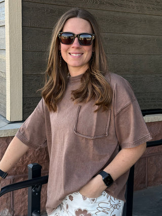 Woman wearing sunglasses and a brown shirt standing against a wooden wall.