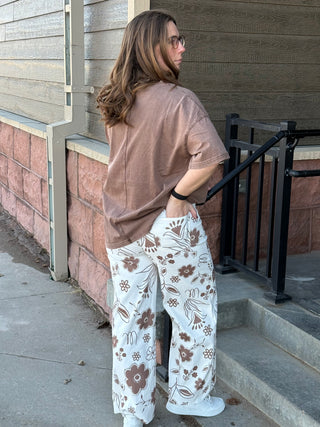 Person wearing a brown shirt and white floral pants standing on a sidewalk.