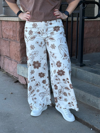 Person wearing white pants with brown floral pattern standing on a sidewalk.