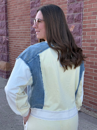 Woman wearing a light-colored jacket with denim patches against a brick wall.