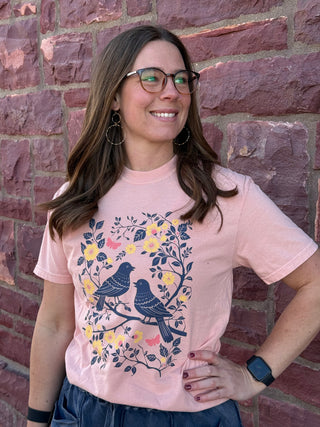 Woman wearing a pink t-shirt with bird design against a brick wall
