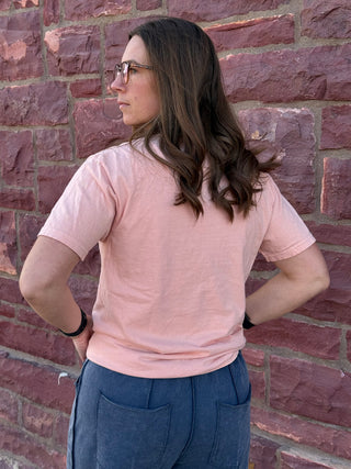 Woman wearing a pink shirt and blue pants standing against a brick wall.