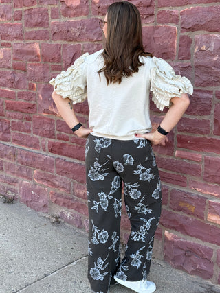 Woman wearing a white blouse with ruffled sleeves and black floral pants against a brick wall.