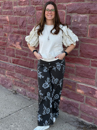 Woman wearing a white blouse with ruffled sleeves and black floral pants against a brick wall.