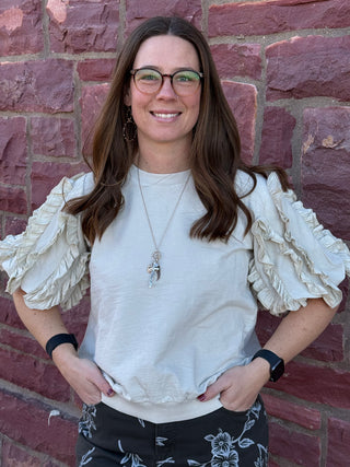 Woman wearing a white blouse with ruffled sleeves against a brick wall.