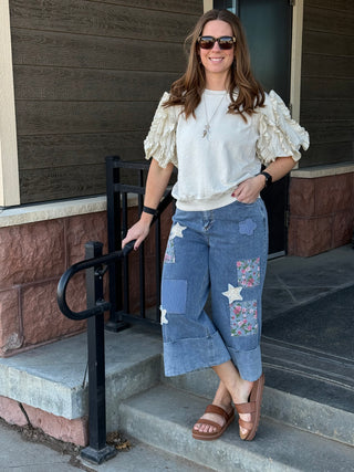 Woman standing on steps wearing a white blouse with ruffled sleeves and blue patchwork pants.