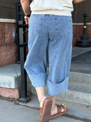 Person wearing blue denim capri pants and brown sandals on a sidewalk.