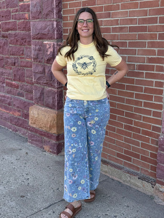 Woman wearing a yellow t-shirt and floral pants standing against a brick wall.