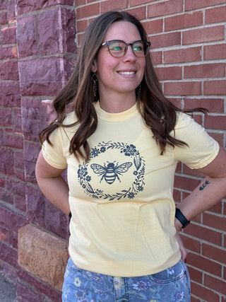 Woman wearing a yellow t-shirt with a bee design against a brick wall.