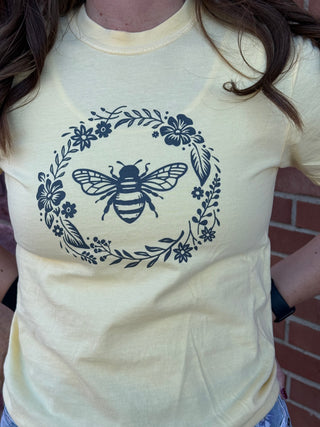 Beige t-shirt with a navy bee and floral design worn by a person against a brick wall.