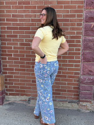 Woman wearing a yellow shirt and floral pants standing against a brick wall.