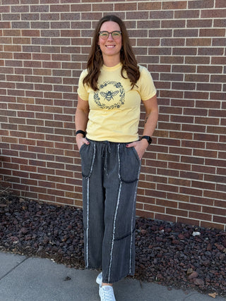 Woman wearing a yellow t-shirt with a graphic design and dark jeans standing in front of a brick wall.