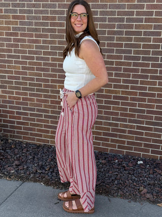 Woman wearing a white sleeveless top and red and white striped pants standing against a brick wall.