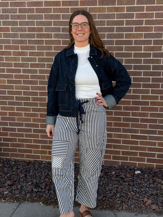 Woman standing against a brick wall wearing a black jacket, white sweater, and striped pants.