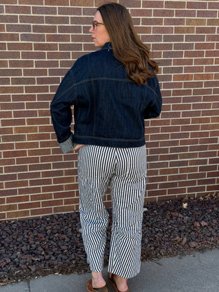 Woman wearing a dark denim jacket and striped pants standing against a brick wall.