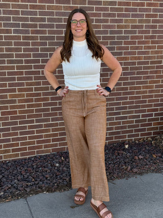 Woman standing against a brick wall wearing a white sleeveless top and brown plaid pants.