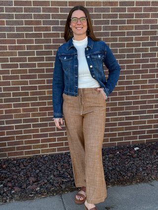 Woman wearing a denim jacket and brown pants standing against a brick wall.