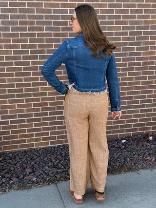 Woman wearing a blue denim jacket and beige plaid pants standing against a brick wall.