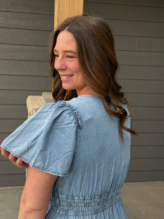 Woman wearing a blue denim dress standing against a gray wall.
