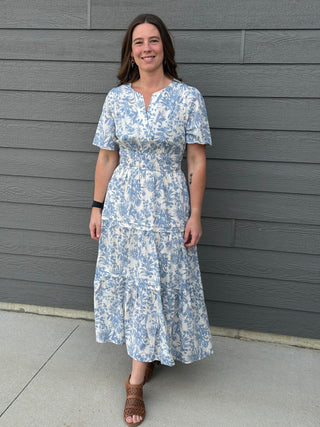Woman wearing a blue floral dress standing against a gray wall.