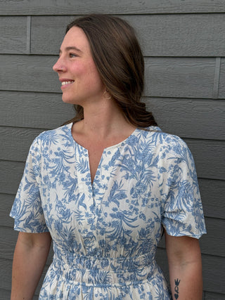 Woman wearing a blue and white floral dress against a gray wooden wall.