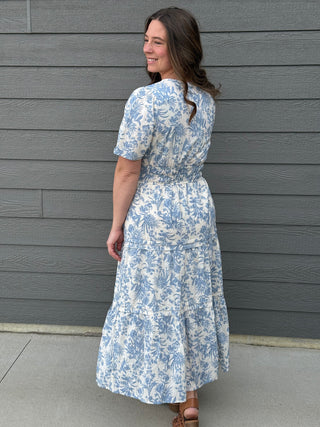 Woman wearing a blue and white floral dress standing against a gray wall.