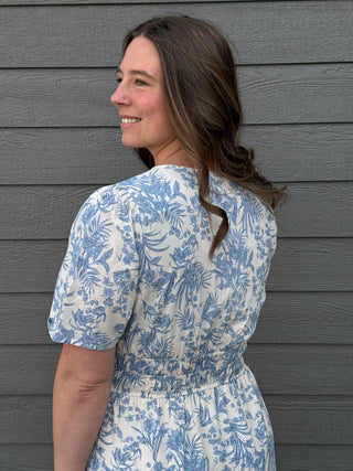 Woman wearing a blue and white floral dress against a gray wooden wall.