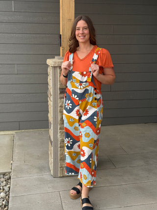 Woman wearing a colorful patterned jumpsuit and orange shirt standing against a gray wall.