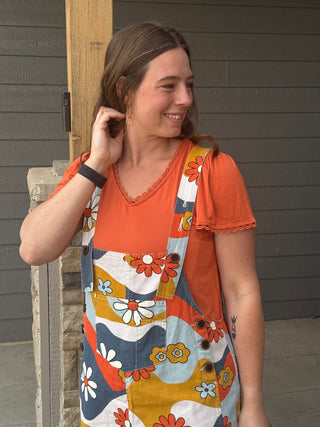 Woman wearing a colorful floral dress over an orange shirt, standing against a gray wall.