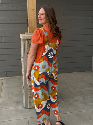 Woman wearing a colorful floral jumpsuit standing against a gray wall.
