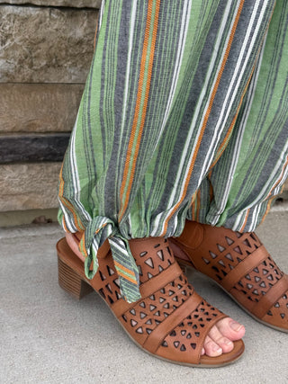 Brown open-toe sandals with cut-out design worn with green striped pants against a stone wall background.