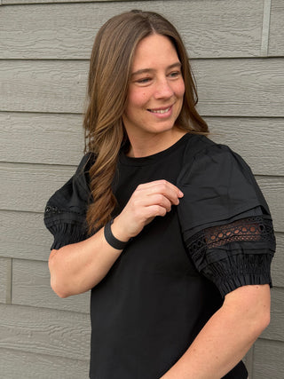 Woman wearing a black top with lace sleeves against a gray wooden wall
